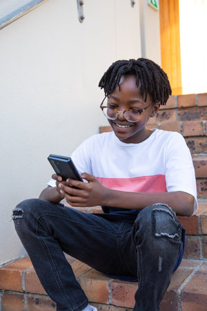 Teenage African American boy sitting on home brick steps wearing eyeglasses holding smartphone....