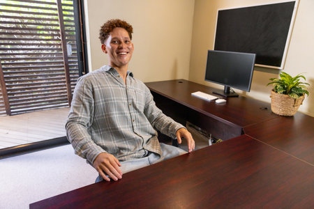 African American man sitting at home office desk smiling at camera with computer monitor and plant....
