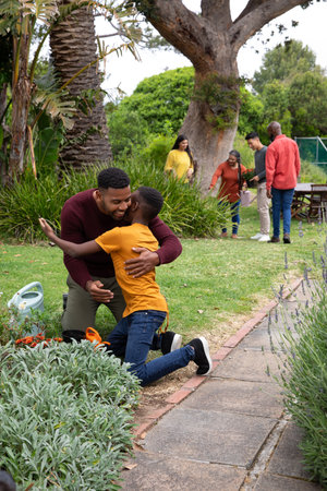 African American family hugging near brick-edged flower bed while using watering can in garden....