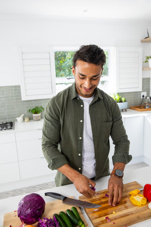 Adult Indian man chopping colorful vegetables on wooden cutting board in bright modern kitchen....