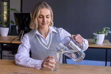 Female office worker pouring water from glass pitcher into tall glass at office desk. Professional, minimalist, contemporary, workspace, hydration, wellness, productivityの写真素材