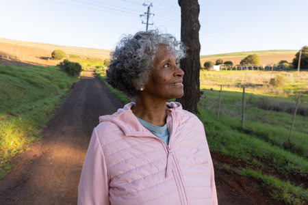 Senior African American woman in pink jacket standing on dirt road gazing at tree, copy space....
