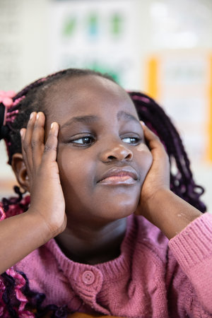 African American girl in pink sweater sitting with face in hands at desk under classroom posters....