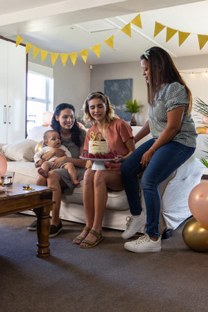 Diverse female friends with baby marking thirtieth birthday in living room with cake and balloons. Celebration, friendship, domestic, modern, cozy, joyful, nurturingの写真素材