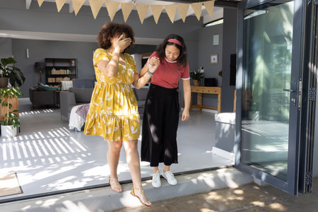 Diverse female friends standing at home door guiding friend through sliding glass door with bunting....