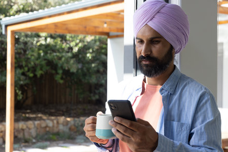 Indian man standing by sliding door at home, holding teal mug and checking smartphone, copy space....