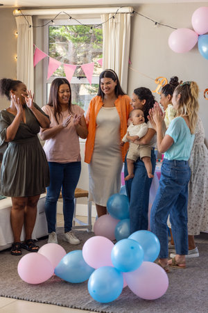 Diverse female friends clapping and celebrating baby shower at home with pastel balloons and banner. Celebration, friendship, joy, pastel, interior, happiness, connectionの写真素材