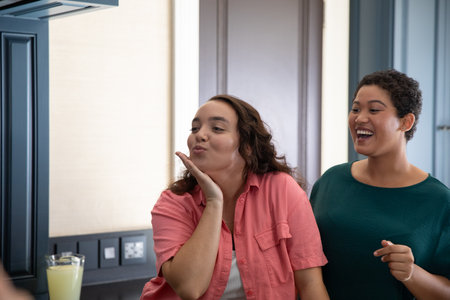 Female friends standing in home kitchen blowing playful kiss toward reflection near juice glass....