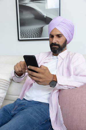Mid adult Indian man wearing turban checking smartphone while sitting on couch in living room....
