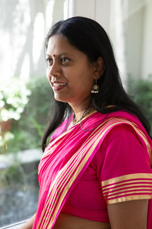 Indian woman wearing pink sari jhumka earrings standing by glass window gazing at garden view....