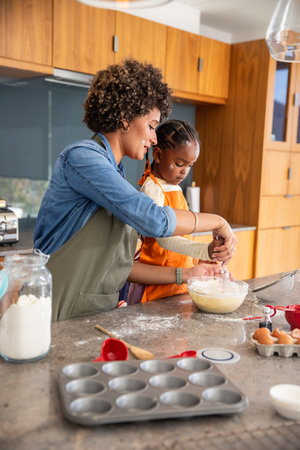 Mother and daughter mixing batter at kitchen countertop using glass bowl whisk and muffin tin....
