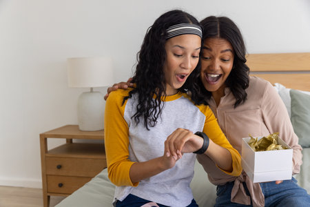 African American mother and teenage daughter opening gift box on bed and admiring wristwatch. Family, surprise, celebration, affection, bonding, gifting, cozyの写真素材