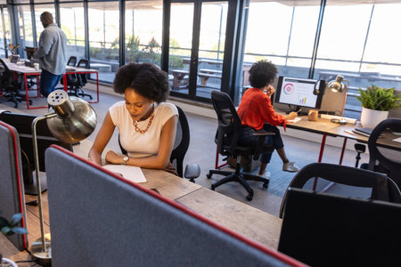 Diverse coworkers writing and viewing charts in open office using notepad, computer and smartphone....