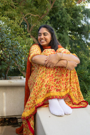 Indian woman sitting on stone ledge on terrace hugging knees in yellow suit and gold bangles....