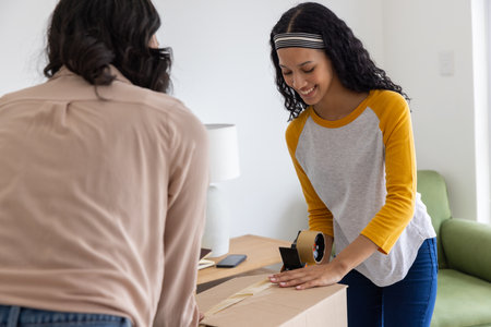 Mother and teenage daughter taping cardboard box with tape dispenser on wooden table in living room. Family, bonding, collaboration, domestic, casual, lifestyle, warmthの写真素材