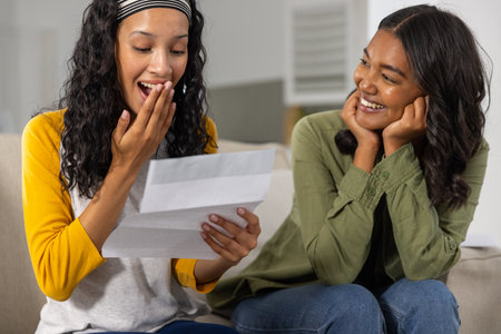 Diverse mother and teenage daughter reacting to folded letter on couch in living room with headband....