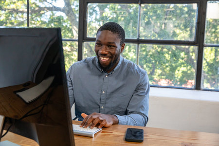 African American man typing on keyboard at office desk by window with monitor and smartphone....
