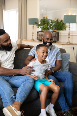 African American family laughing while touching child's shoulders on grey sofa in living room....