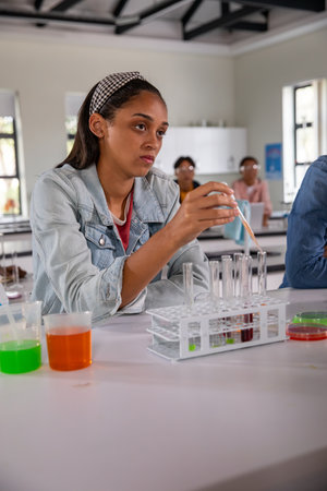 Diverse teenage classmates using pipette and transferring liquids into test tube rack on lab bench....