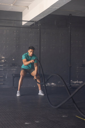 Male athlete performing battle rope exercise while sitting on bench in minimalist gym. Strength, fitness, training, exercise, endurance, sport, athleticism