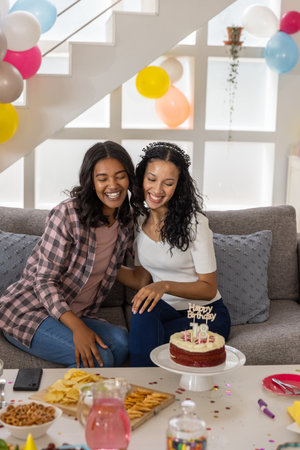 Female friends celebrating 18th birthday in living room with red velvet cake and pastel balloons....