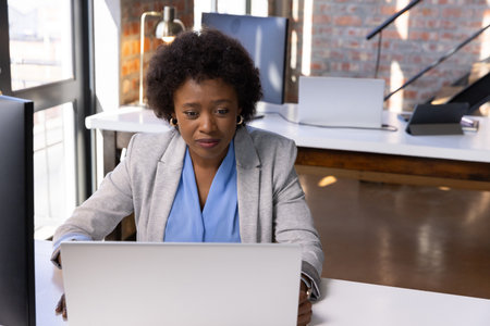 African American woman in blazer typing on laptop at office workstation with lamp, microphone stand....