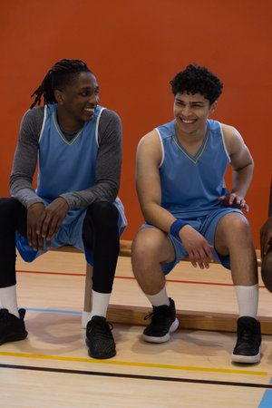 Diverse male teammates wearing blue jerseys sitting on bench on basketball court holding basketball....
