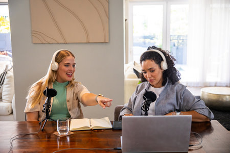 Diverse female co-hosts recording podcast in living room at wooden table with laptop microphones....