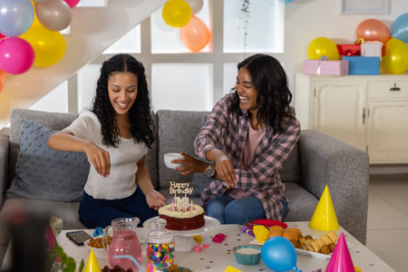 Diverse female friends celebrating birthday on couch in living room with cake, balloons, party hats....