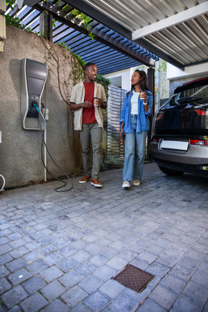 African American couple walking under carport holding coffee cups near EV charging station. Modern,...