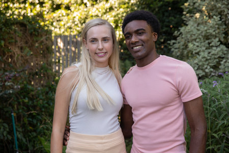 White woman and African American man couple standing in sunny garden wearing cream top pink t-shirt. Romantic, outdoor, vibrant, natural, affectionate, leisure, pastoralの写真素材