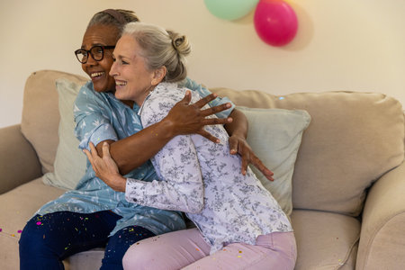 Senior female friends hugging on beige sofa in living room with pastel balloons and confetti. Warmth, companionship, celebration, relaxation, elegance, comfort, leisureの写真素材