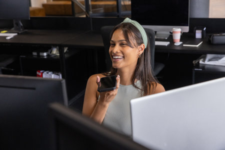 Asian woman smiling and holding smartphone while speaking at modern office desk with dual monitors....