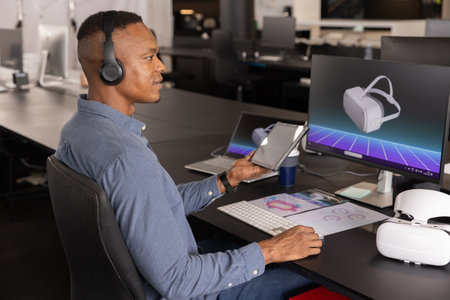 Mid adult African American man holding tablet, viewing VR headset unit at office desk, copy space....