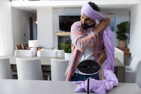 Mid adult Indian man tying lavender turban cloth on kitchen island beside vanity mirror, copy space....