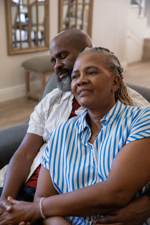 African American couple sitting on gray couch in living room with side table and staircase railing. Romantic, companionship, comfort, serenity, homey, cozy, affectionateの写真素材