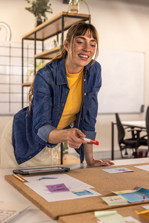 Female designer leaning over cork board in office marking sketches and sticky notes with red marker....