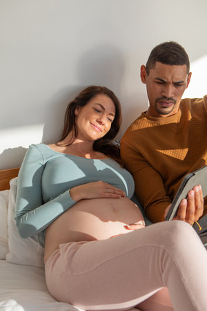 Diverse couple sitting on bed in bedroom against headboard cradling bare pregnant belly with tablet....
