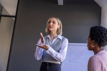Diverse female coworkers presenting data on flip chart with colorful line graph in conference room. Collaboration, professional, innovation, teamwork, communication, strategy, modernの写真素材