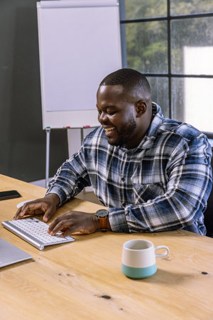 African American man typing on wireless keyboard at desk with laptop, mug, smartphone, flipchart. Professional, technology, workplace, modern, productivity, corporate, communicationの写真素材