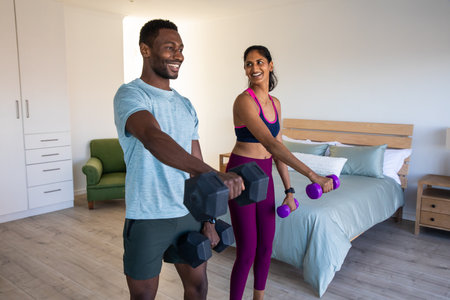 Diverse couple holding black and purple dumbbells while working out in bedroom at home. Fitness,...