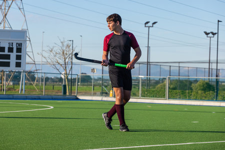 Teenage boy gripping hockey stick with neon green handle gazing left on turf pitch near scoreboard. Athlete, outdoor, vibrant, competitive, training, sport, sportswearの写真素材