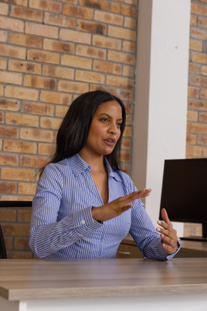 Female office professional gesturing while presenting ideas at office desk with computer monitor. Corporate, teamwork, innovation, workspace, industry, collaboration, modernの写真素材