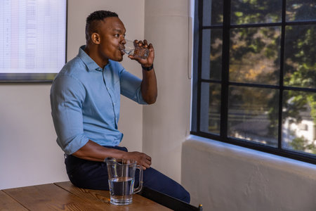 African American man drinking water from glass in conference room with pitcher and screen. Corporate, professionalism, refreshment, modernity, workspace, productivity, wellnessの写真素材