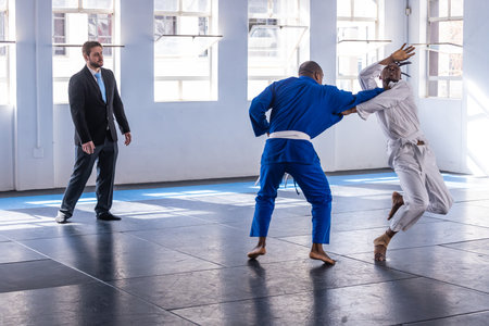 Diverse male sparring fighters wearing gis exchanging grips on mats in dojo with referee watching....