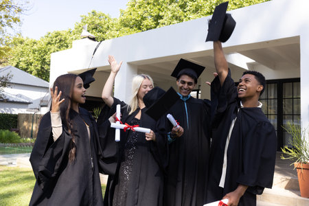 Diverse classmates celebrating on campus lawn wearing black gowns tossing caps and holding diplomas. Commencement, achievement, jubilation, camaraderie, outdoor, modern, vibrantの写真素材
