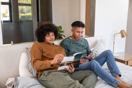 Diverse male friends sitting on bed reading open book and scrolling smartphone in modern bedroom....