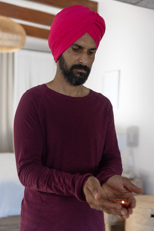 Indian man holding small oil lamp in softly lit bedroom wearing red turban and maroon shirt....
