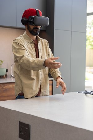 Indian man using VR headset with headphones at kitchen countertop, manipulating virtual interface....