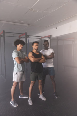 Diverse male workout partners standing with arms crossed in gym before pull-up and squat rack....